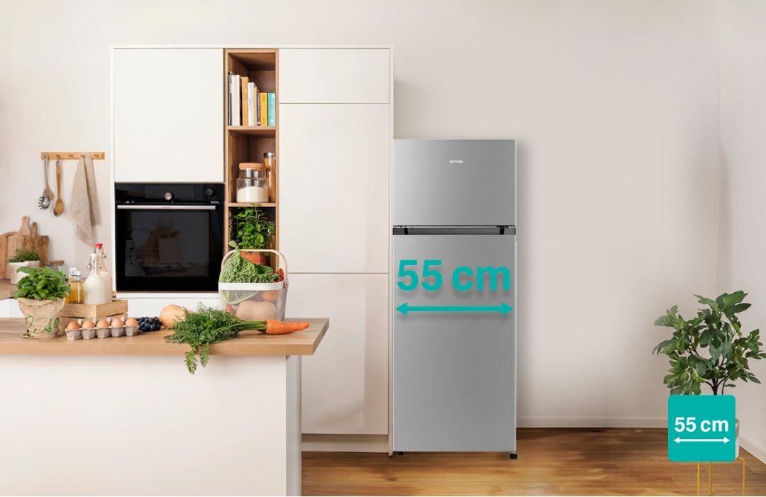 W kuchni znajduje się lodówka w kolorze inox z napisem \'55 cm\' wskazującym szerokość. Obok lodówki jest zabudowa kuchenna z piekarnikiem i półkami, a na blacie leżą różne produkty spożywcze.