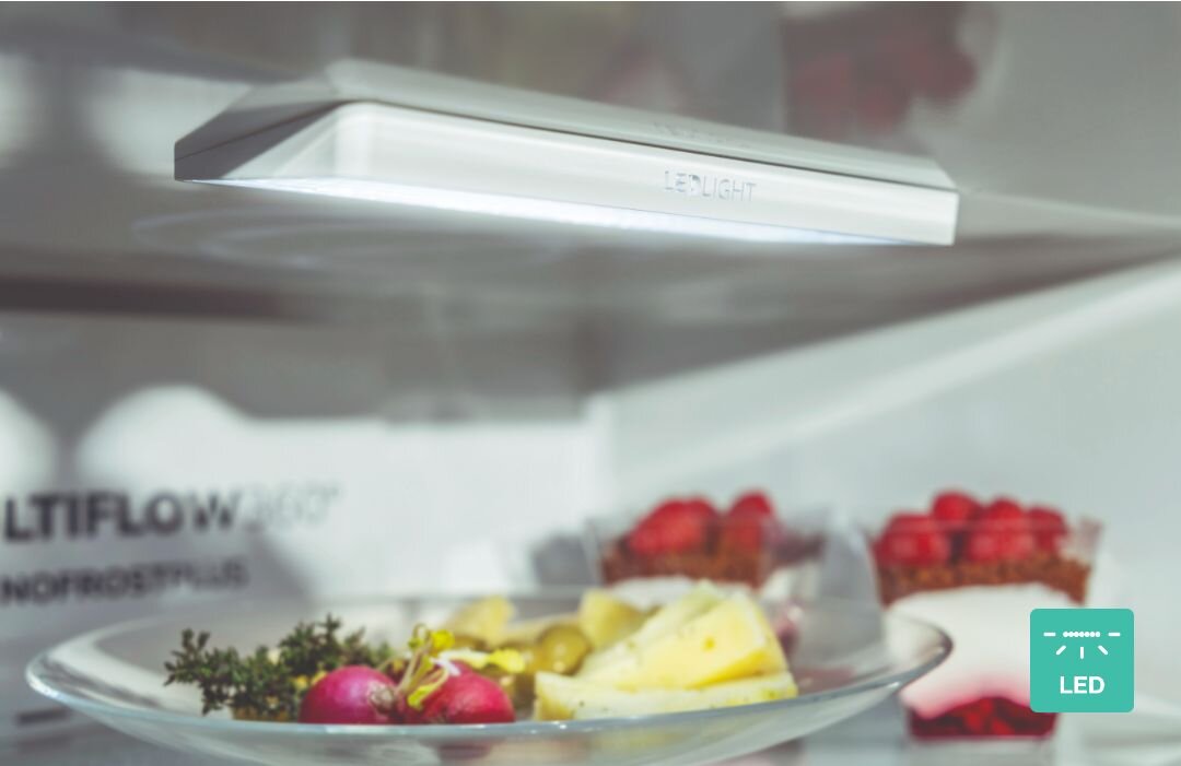 Wnętrze lodówki z oświetleniem LED, na półce znajduje się talerz z owocami i warzywami. Widoczny tekst: \'LED LIGHT\', \'MULTIFLOW 360°\', \'NOFROST PLUS\'.