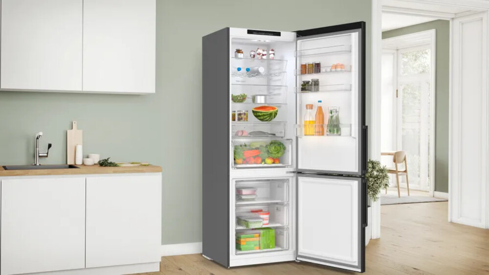 Otwarta lodówka z widocznymi półkami wypełnionymi różnymi produktami spożywczymi, takimi jak warzywa, napoje i słoiki. Wnętrze jest jasno oświetlone, a lodówka stoi w nowoczesnej kuchni z drewnianą podłogą.
