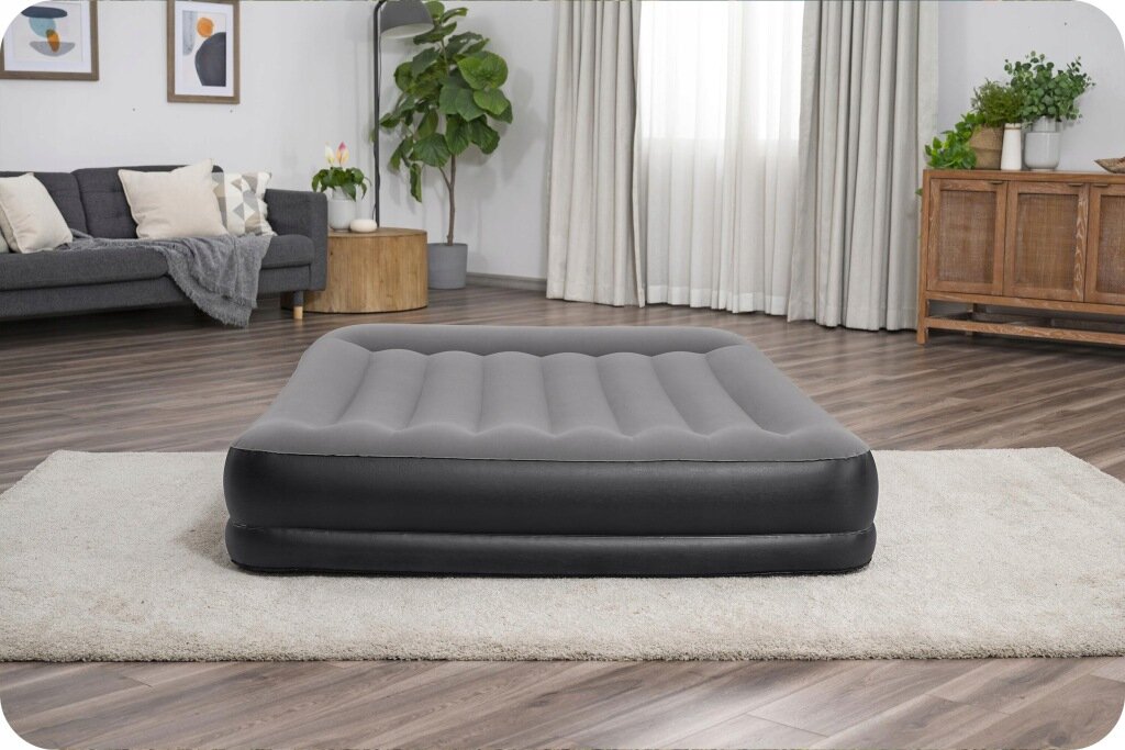 Na obrazie znajduje się szary, dmuchany materac dwuosobowy umieszczony na jasnym dywanie w salonie. W tle widoczna jest sofa, rośliny doniczkowe i zasłony na oknie.