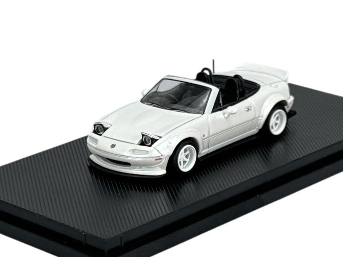 Model samochodu w skali 1:64 przedstawiający białą Mazdę MX-5 Miata z otwartym dachem, umieszczony na czarnej podstawce.