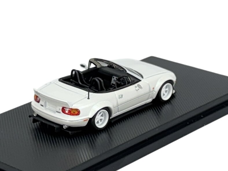 Model samochodu w skali 1:64 przedstawiający białą Mazdę MX-5 Miata z otwartym dachem, umieszczony na czarnej podstawce. Samochód ma białe felgi i widoczne detale wnętrza.