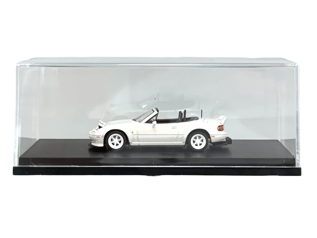 Model samochodu w skali 1:64 przedstawiający białą Mazdę MX-5 Miata, umieszczony w przezroczystej gablocie na czarnej podstawce. Samochód ma otwarty dach i widoczne szczegóły, takie jak felgi i lusterka.