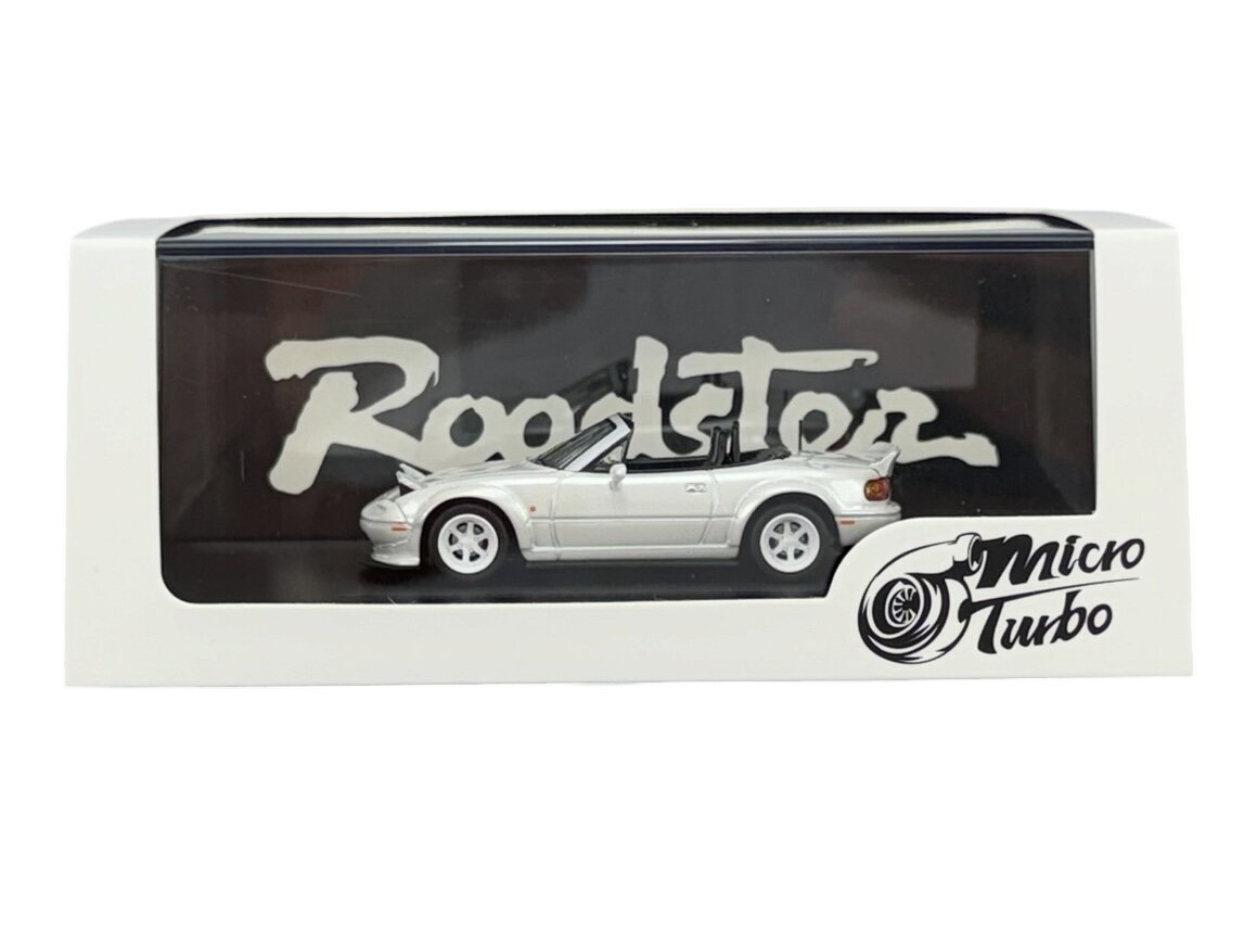 Model samochodu w skali 1:64 w kolorze białym znajduje się w przezroczystym opakowaniu z napisem \'Roadster\'. Na opakowaniu widoczne jest logo \'Micro Turbo\'.