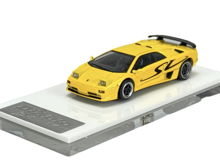 Żółty model samochodu sportowego w skali 1:64 z napisem \'SV\' na boku, umieszczony na białej podstawce z metalową tabliczką. Samochód ma czarne detale i widoczne felgi.