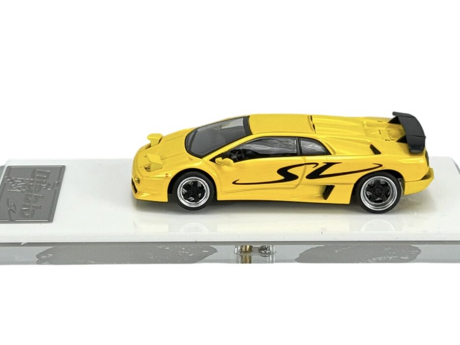 Żółty model samochodu sportowego z czarnymi detalami i napisem \'SV\' na boku, umieszczony na białej podstawce. Obok znajduje się metalowa tabliczka z nieczytelnym napisem.