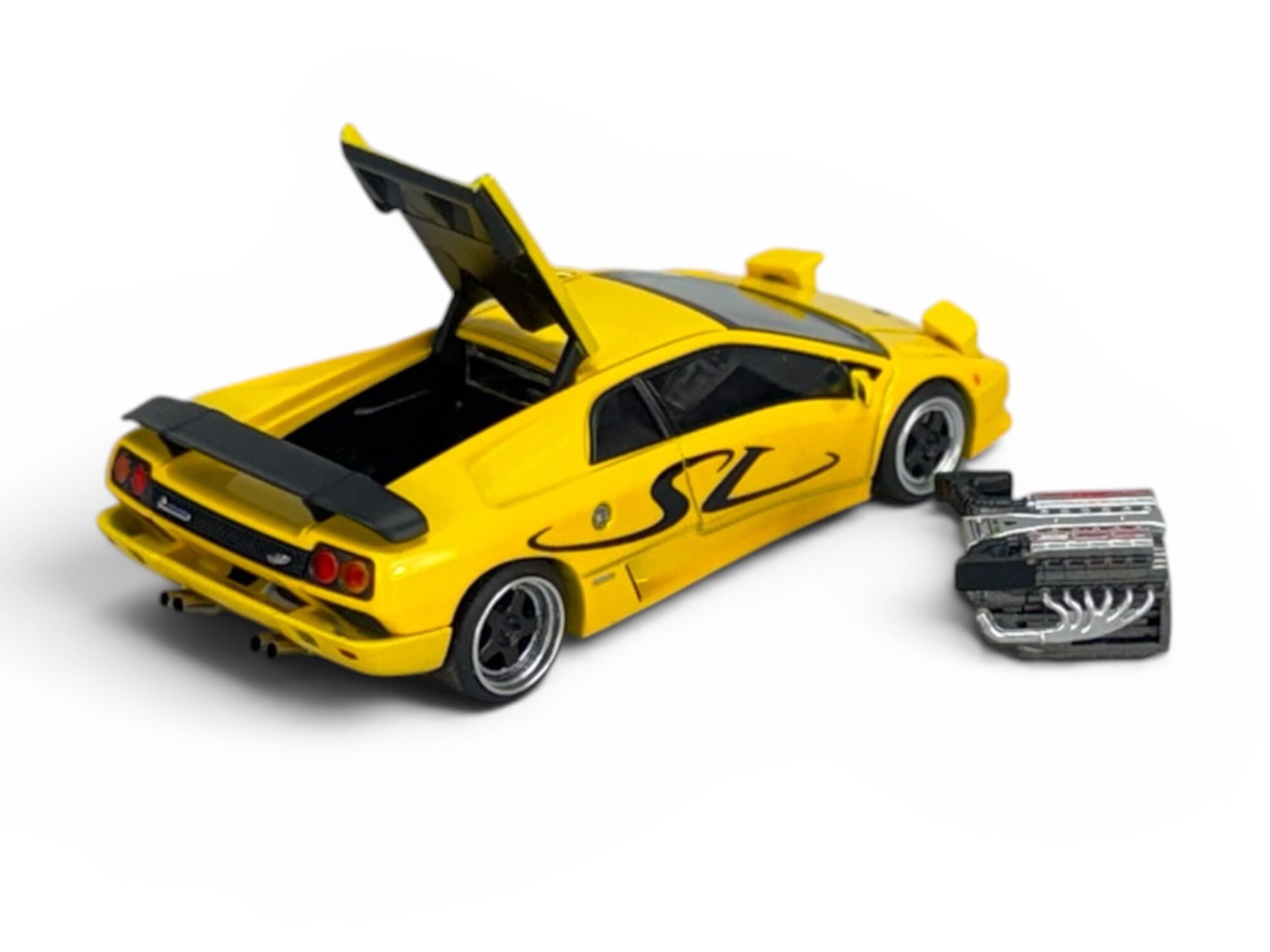 Żółty model samochodu sportowego z otwartym tylnym bagażnikiem i widocznym napisem \'SV\' na boku. Obok modelu znajduje się miniaturowy silnik.