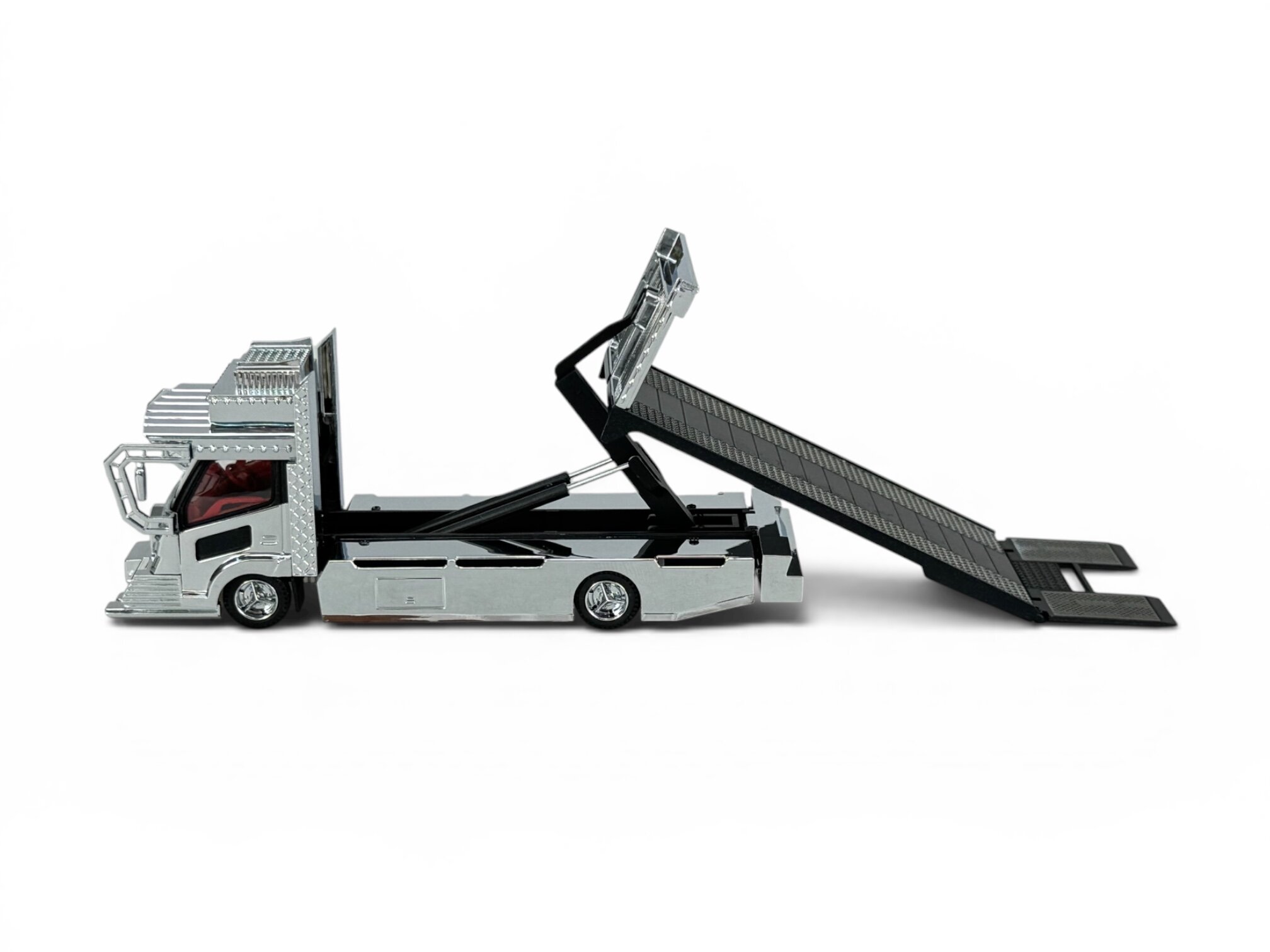 Model metalowej lawety w skali 1:64 z podnoszoną rampą, w kolorze srebrnym z czarnymi elementami. Kabina kierowcy jest szczegółowo wykonana z widocznymi czerwonymi siedzeniami.