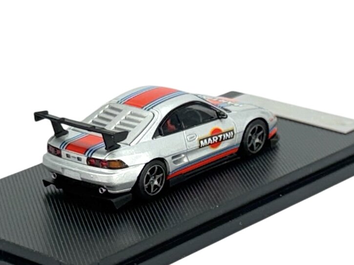 Model samochodu w skali 1:64 przedstawia srebrny pojazd z czerwono-niebieskimi pasami i napisem \'MARTINI\' na boku. Samochód ma duży tylny spojler i stoi na czarnej podstawce.
