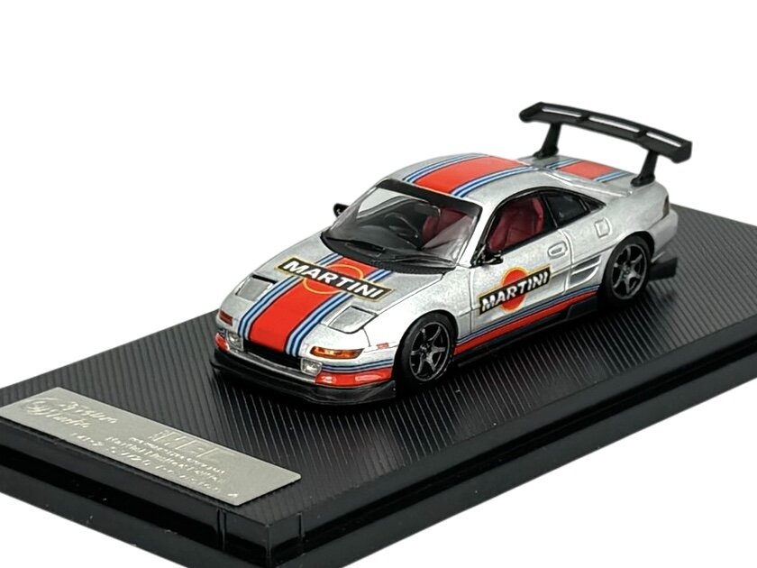 Model samochodu w skali 1:64, srebrny z czerwono-niebieskimi pasami i logo \'MARTINI\' na masce i bokach, umieszczony na czarnej podstawce.