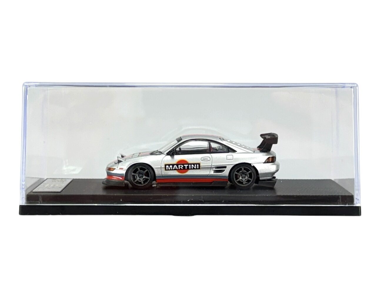Model samochodu w skali 1:64 w kolorze srebrnym z napisem \'MARTINI\' na boku, umieszczony w przezroczystej gablocie na czarnej podstawce.