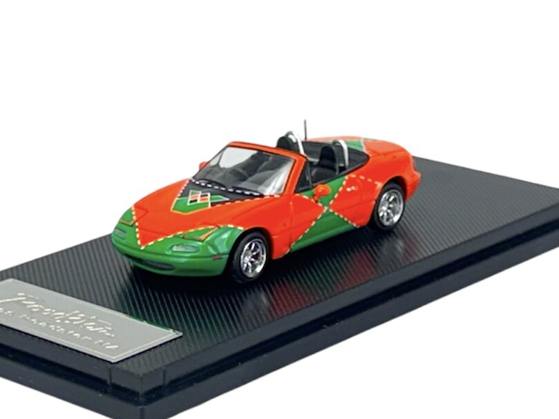Model samochodu w skali 1:64 przedstawia sportowy kabriolet w kolorach pomarańczowym i zielonym z geometrycznymi wzorami. Pojazd umieszczony jest na czarnej podstawce z napisem \'media expert\'.