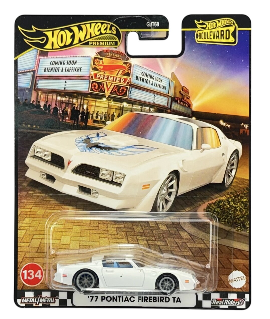 Opakowanie z modelem samochodu \'77 Pontiac Firebird TA w kolorze białym, z logo Hot Wheels Premium i oznaczeniem 134. W tle widoczny jest budynek kina z napisami \'COMING SOON\' i \'BIENTÔT À L\'AFFICHE\'.