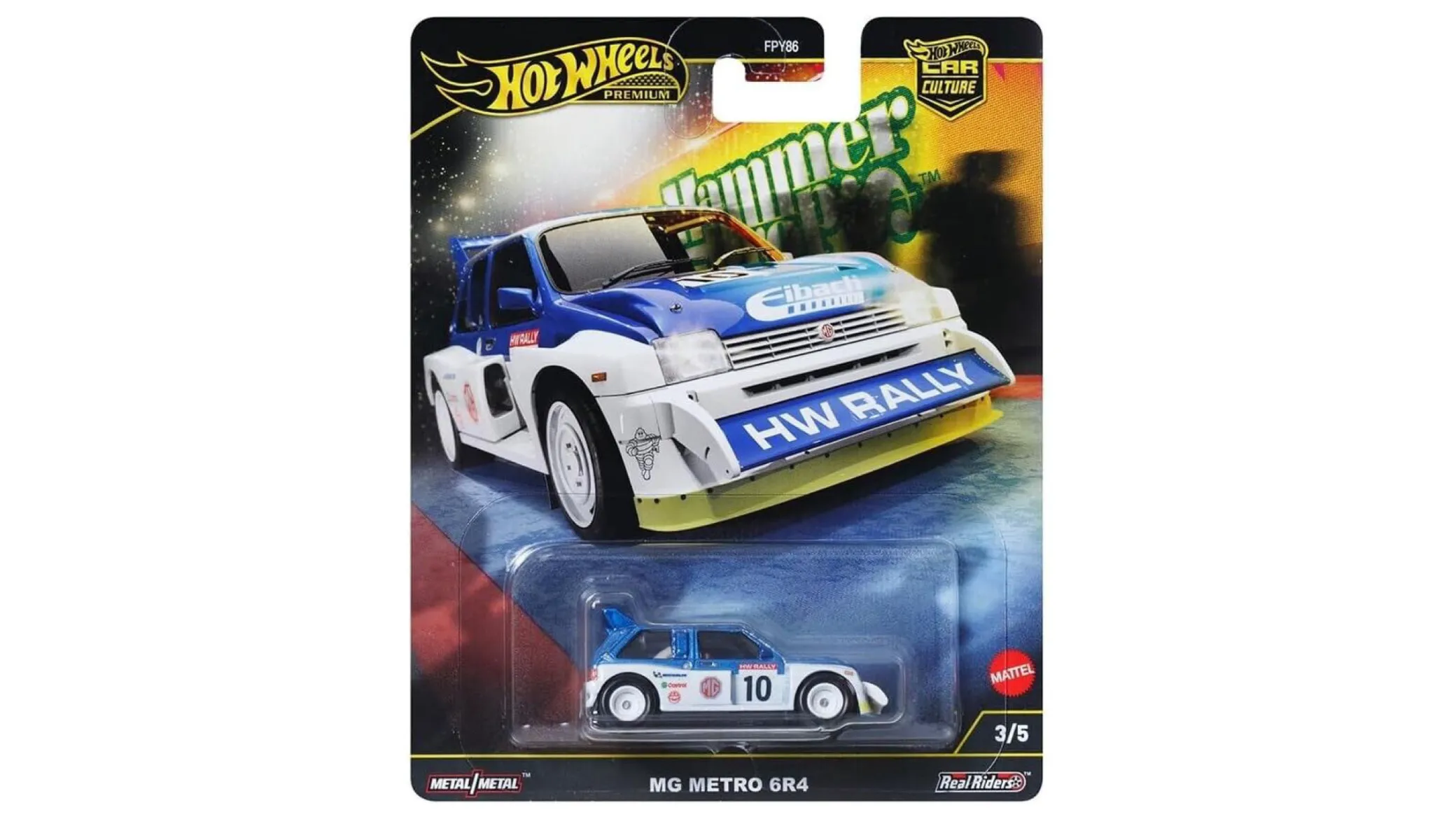 Opakowanie z modelem samochodu Hot Wheels Premium, przedstawiające biało-niebieski samochód rajdowy z napisem \'HW RALLY\' na przednim zderzaku. Widoczne są napisy: \'Hammer Drops\', \'MG METRO 6R4\', \'Metal/Metal\', \'Real Riders\', oraz logo Mattel.