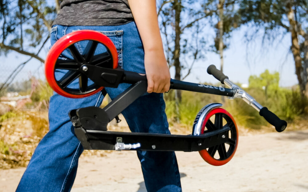 Hulajnoga RAZOR Carbon Lux 13073003 Czarny osoba niesie hulajnogę w ręku lekka aluminiowa rączka wygodny chwyt i trwałość funkcja składania łatwe przechowywanie i przenoszenie waga 4,53 kg