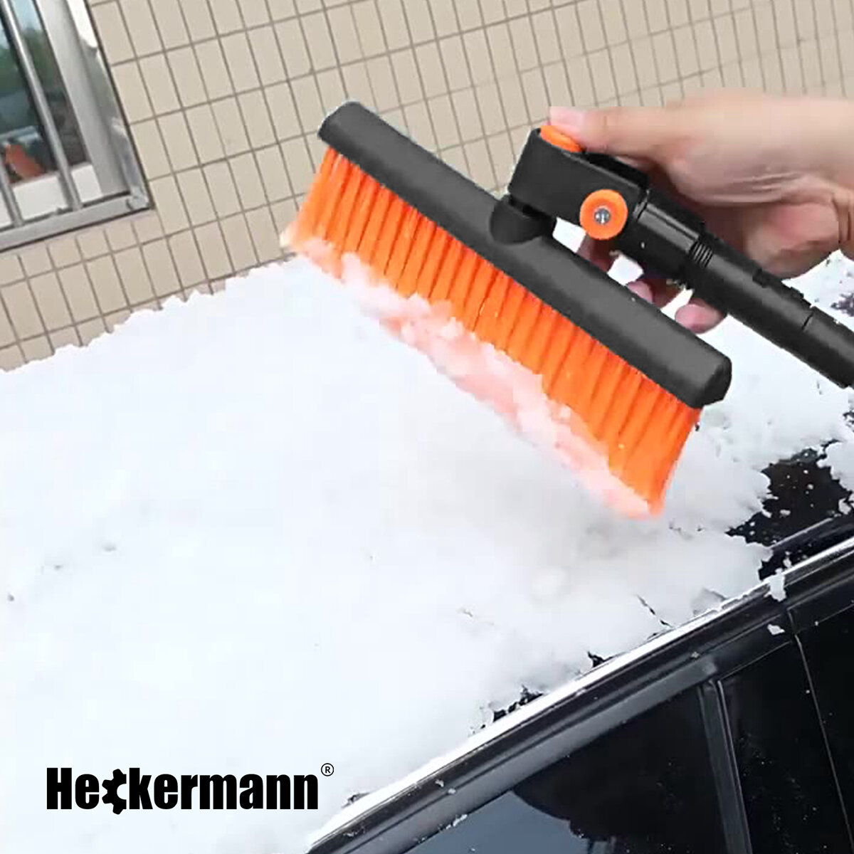Osoba trzyma szczotkę do odśnieżania z pomarańczowym włosiem, usuwającą śnieg z powierzchni samochodu. Widoczne jest logo \'Heckermann\'.