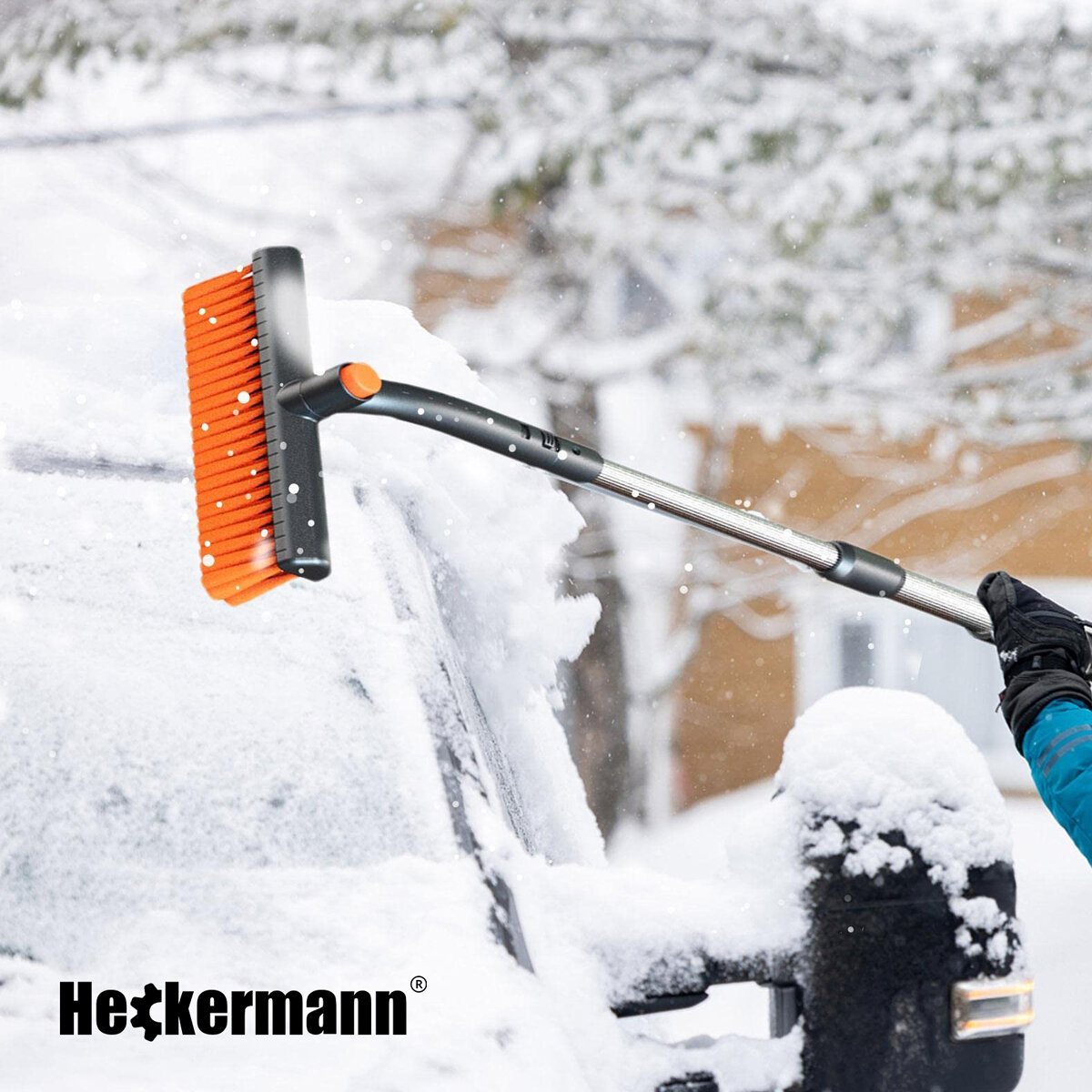 Osoba używa pomarańczowej szczotki do odśnieżania samochodu pokrytego śniegiem. Widoczne jest logo \'Heckermann\'.