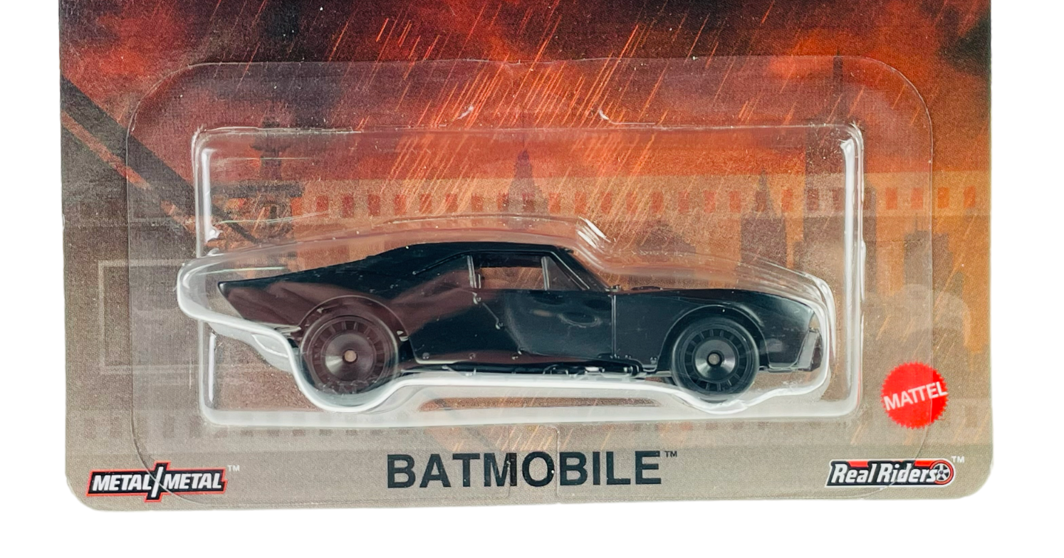Czarny model samochodu Batmobile w opakowaniu z przezroczystego plastiku, na tle grafiki z motywem miasta. Widoczne logo \'Mattel\', \'Metal/Metal\' oraz \'Real Riders\'.
