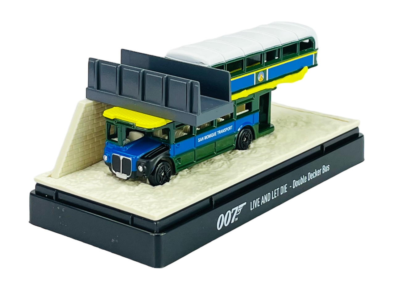 Model metalowego autobusu piętrowego w kolorach zielonym, niebieskim i żółtym, umieszczony na podstawce z napisem \'007 LIVE AND LET DIE - Double Decker Bus\'. Na boku autobusu widoczny jest napis \'SAN MONIQUE TRANSPORT\'.