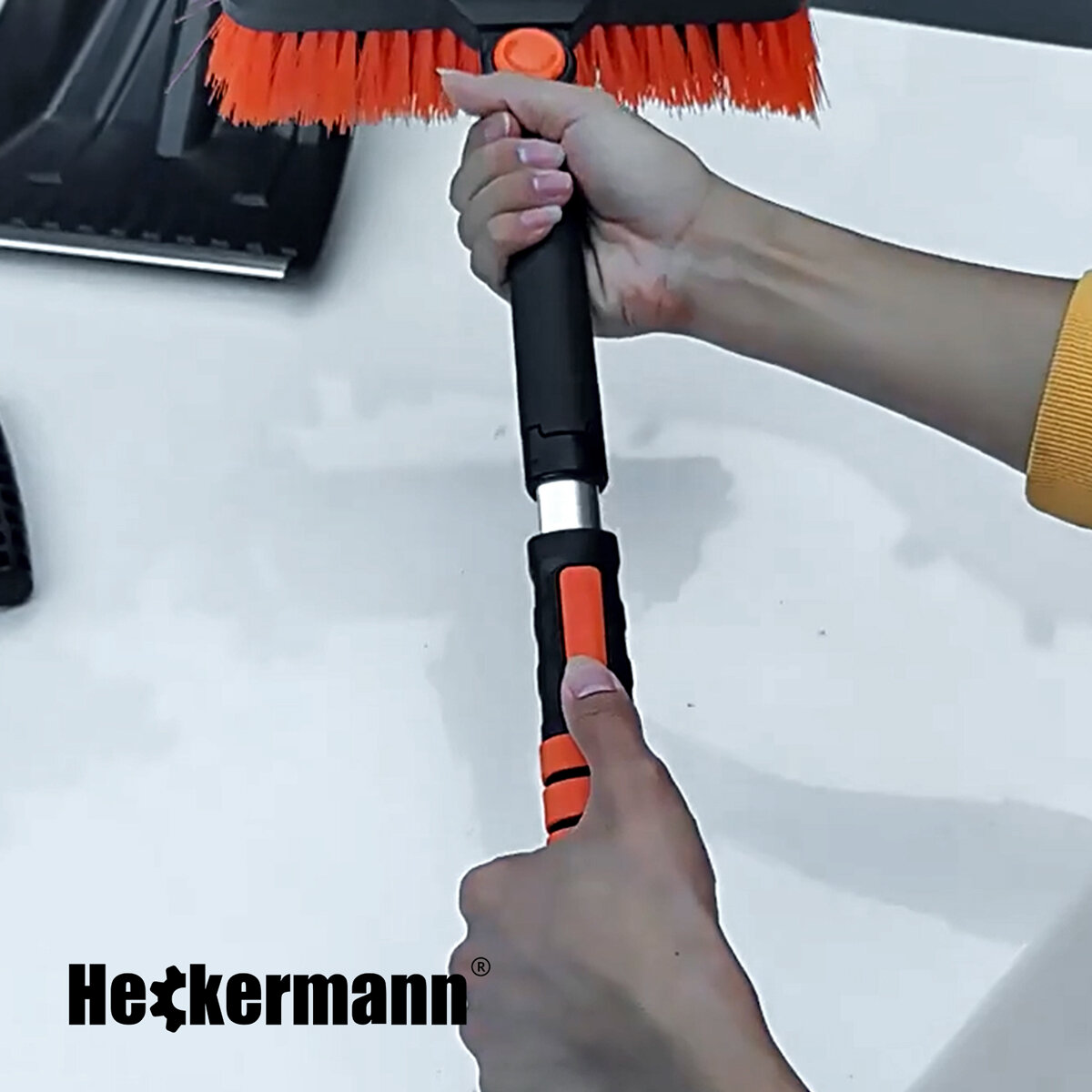 Osoba trzyma narzędzie z pomarańczowymi włosiami i czarną rączką. Widoczne jest logo \'Heckermann\'.