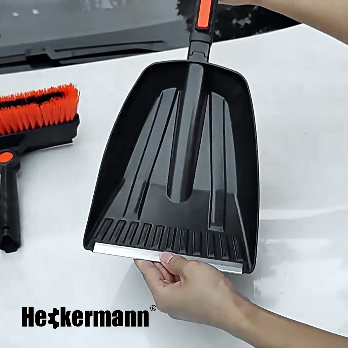 Osoba trzyma czarną łopatkę z pomarańczowym uchwytem, obok widoczna jest szczotka z pomarańczowym włosiem. Na obrazie znajduje się logo \'Heckermann\'.