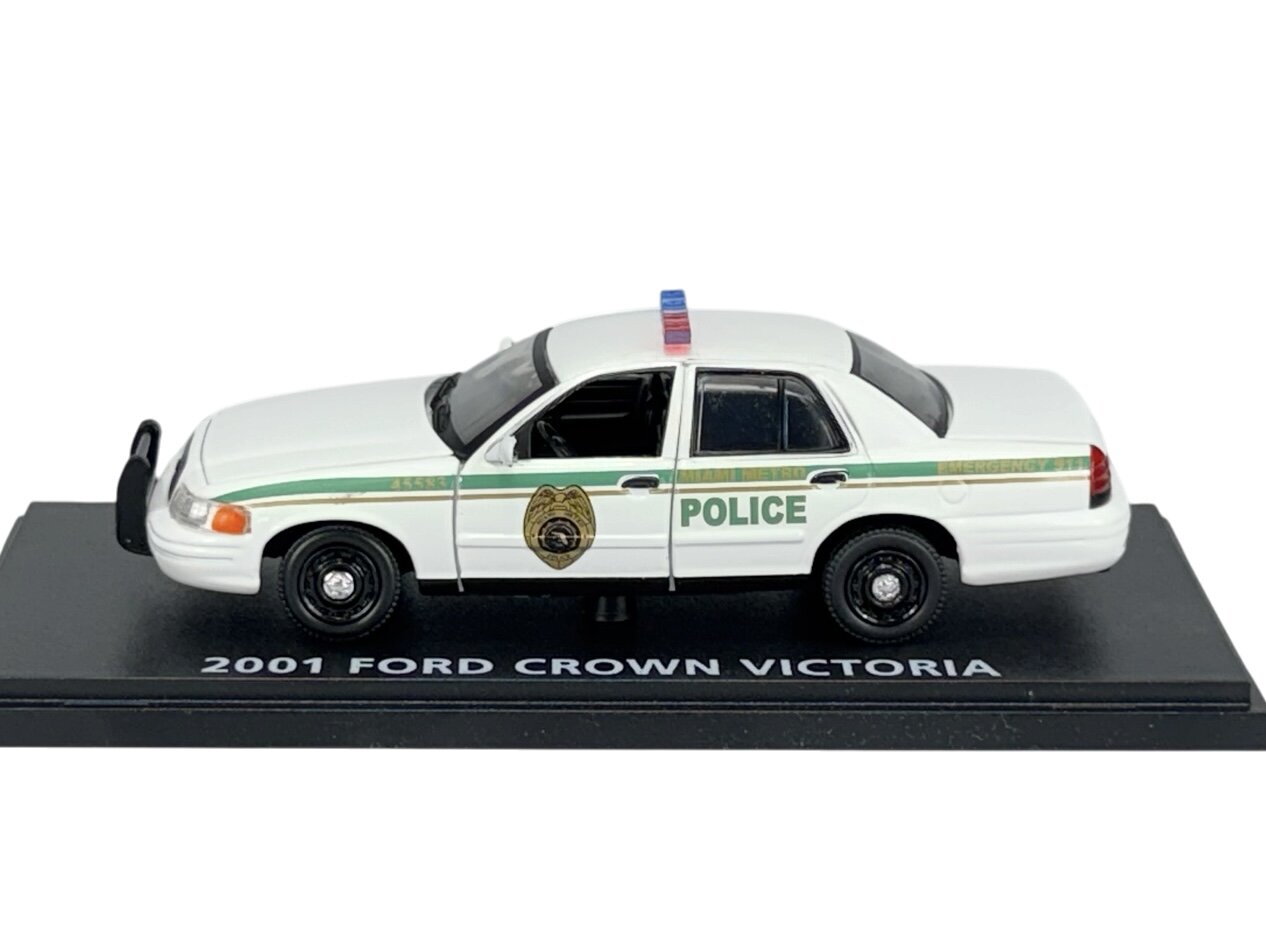 Model policyjnego samochodu Ford Crown Victoria z 2001 roku w kolorze białym z zielonymi detalami i napisem \'POLICE\' na boku. Samochód umieszczony jest na podstawce z napisem \'2001 FORD CROWN VICTORIA\'.