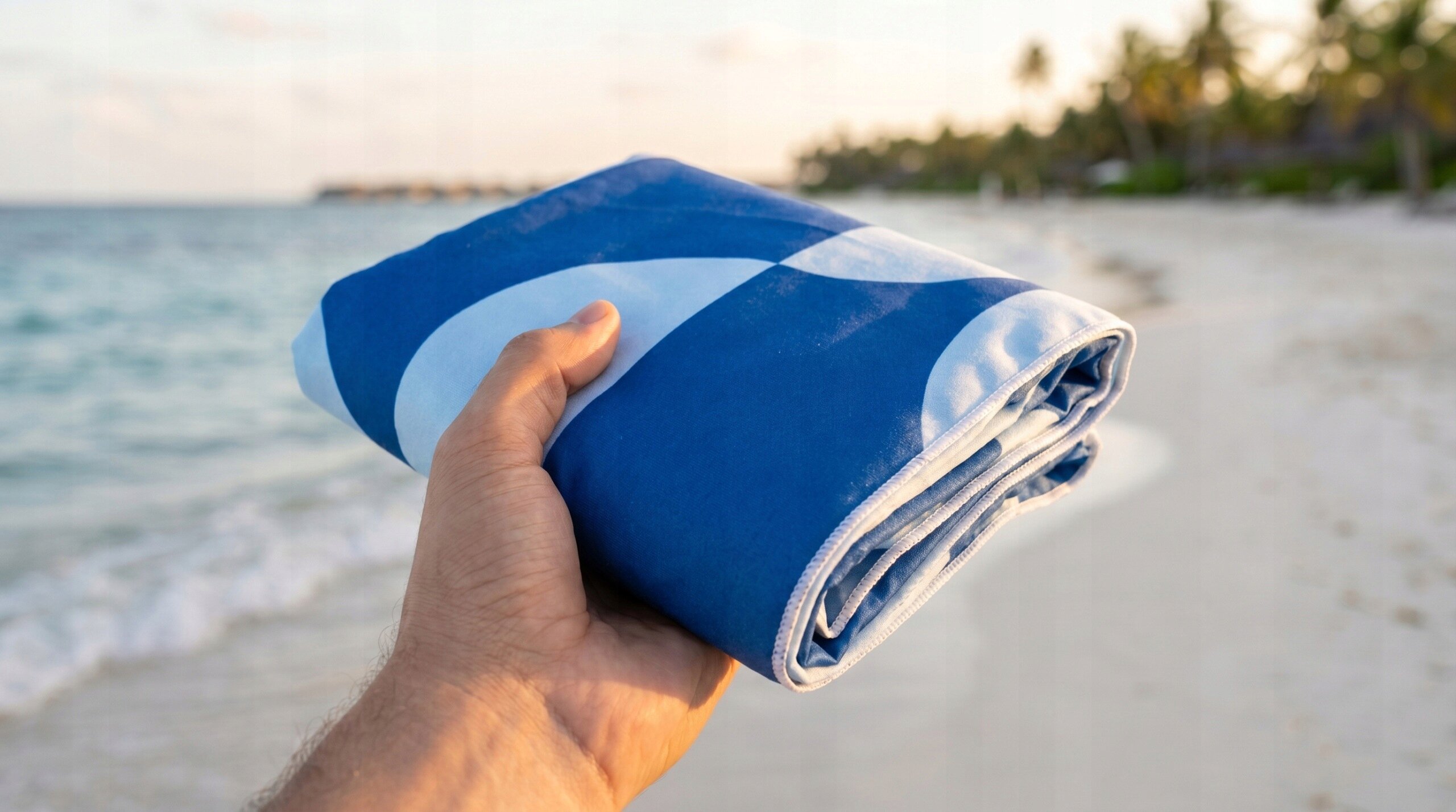 Osoba trzyma złożony, niebieski ręcznik z białymi wzorami na tle plaży z palmami i morzem.
