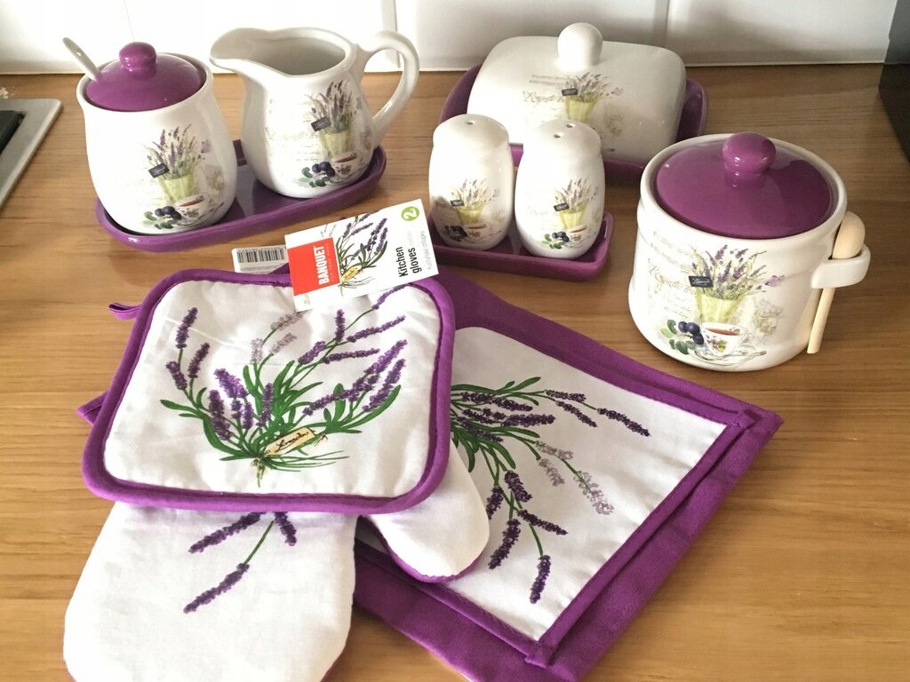 Na drewnianym blacie znajduje się zestaw ceramiczny z motywem lawendy, w skład którego wchodzą mlecznik, cukierniczka, solniczka, pieprzniczka, maselniczka oraz tekstylia kuchenne z fioletowym obramowaniem. Widoczna jest etykieta z napisem \'media expert\'.
