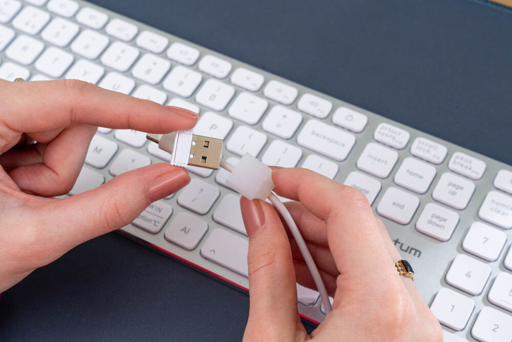 Osoba trzyma w rękach kabel USB oraz adapter, na tle białej klawiatury z widocznym napisem \'XQUANTUM\'.