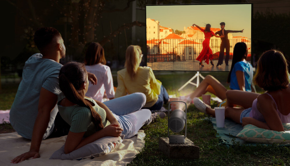 Grupa osób siedzi na trawie, oglądając film wyświetlany na ekranie z projektora. Projektor stoi na ziemi, a na ekranie widać parę tańczącą na tle miasta.