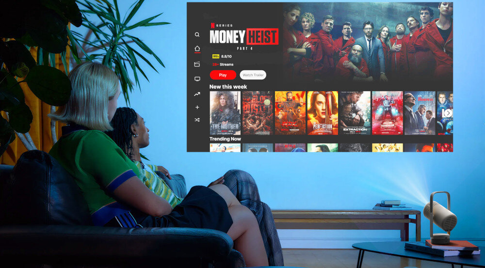 Dwie osoby siedzą na kanapie, oglądając projekcję na ścianie z interfejsem platformy streamingowej, na której widoczny jest serial \'Money Heist\'. Obok stoi projektor, a na ekranie widać także inne propozycje filmowe i serialowe.