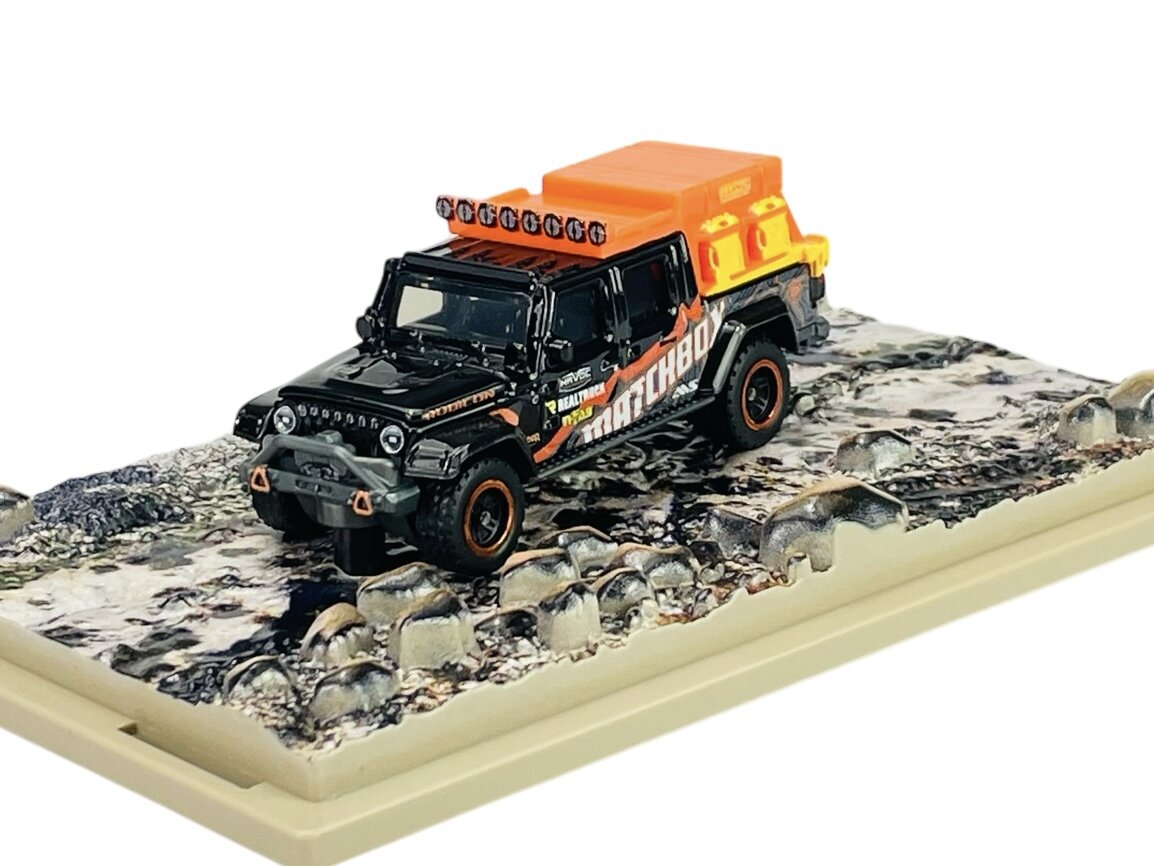 Metalowy model samochodu terenowego w czarnym kolorze z pomarańczowymi akcentami i napisem \'MATCHBOX\' na boku, umieszczony na podstawce imitującej skalisty teren.