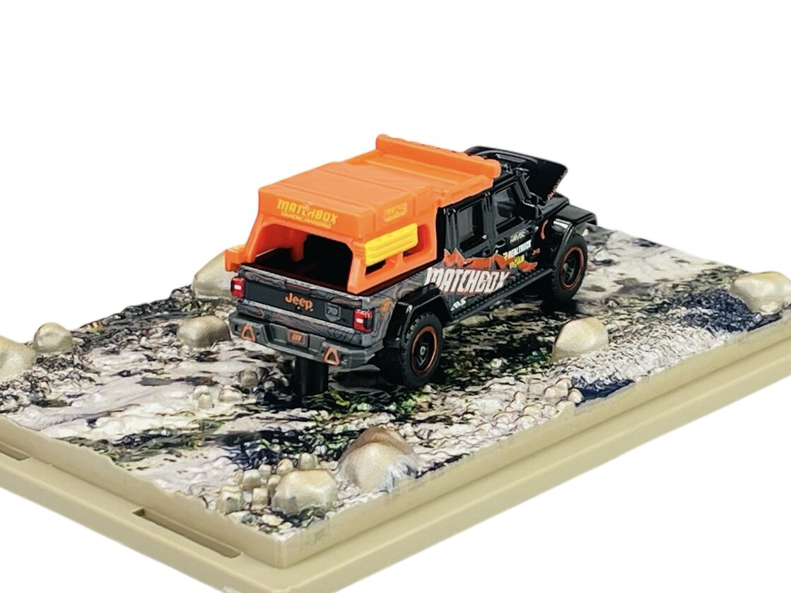 Model metalowego samochodu terenowego z pomarańczowym dachem i napisami \'Matchbox\' oraz \'Jeep\' na tylnej części. Samochód stoi na podstawce imitującej kamienistą drogę.