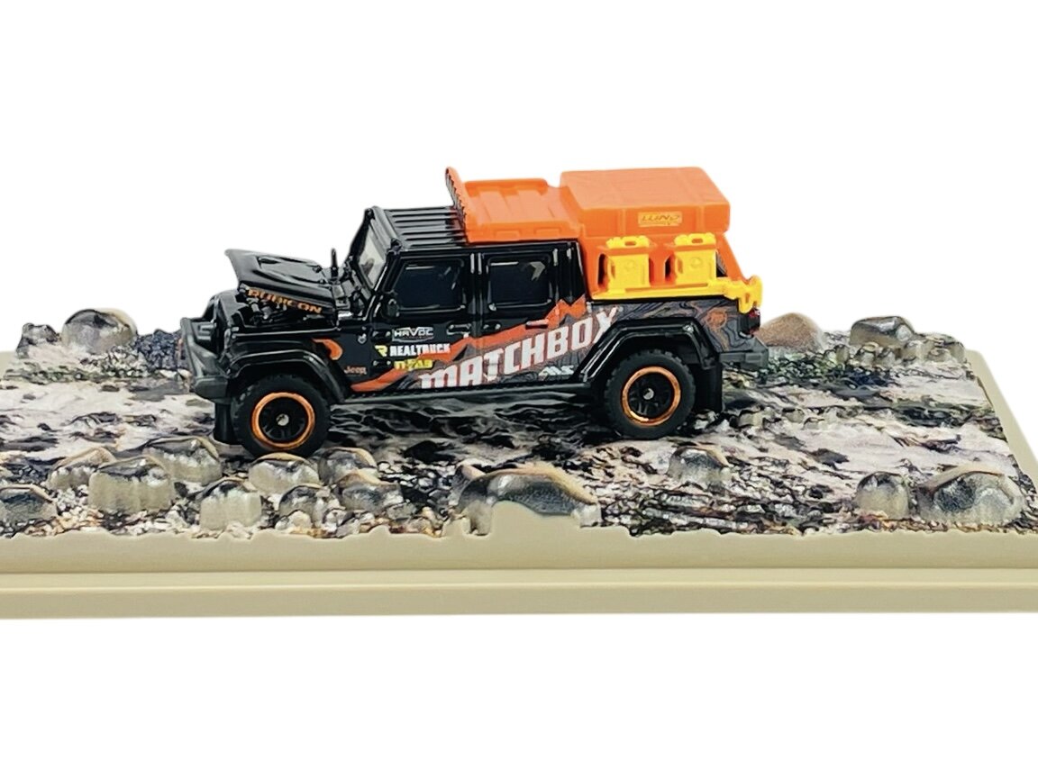 Metalowy model Jeepa Gladiatora w czarnym kolorze z pomarańczowymi akcentami i napisem \'MATCHBOX\' na boku. Samochód stoi na podstawce imitującej teren skalisty.
