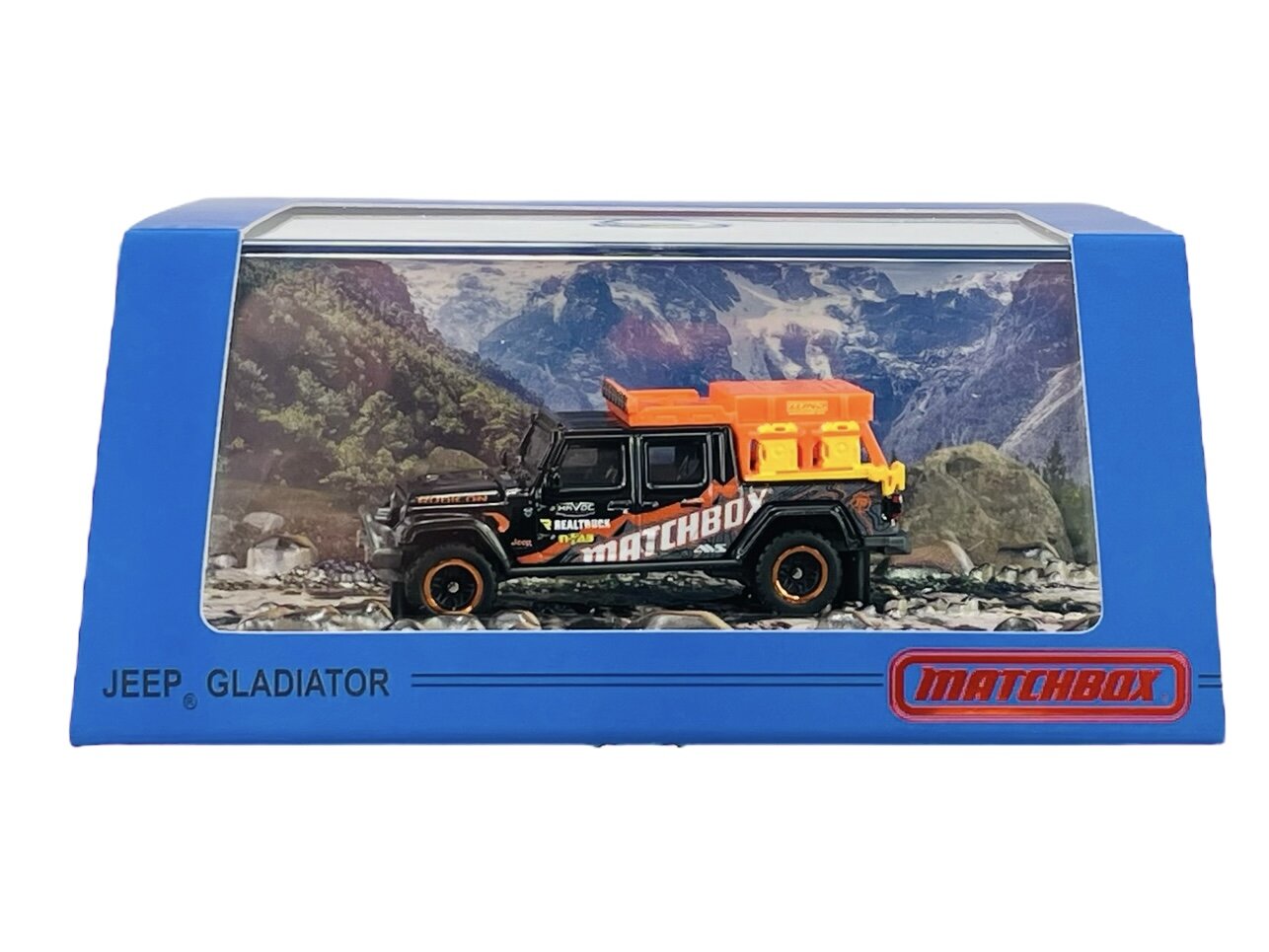 Model metalowego samochodu Jeep Gladiator w opakowaniu z przezroczystym frontem, na tle górskiego krajobrazu. Na opakowaniu widoczne są napisy: \'JEEP GLADIATOR\' i \'MATCHBOX\'.