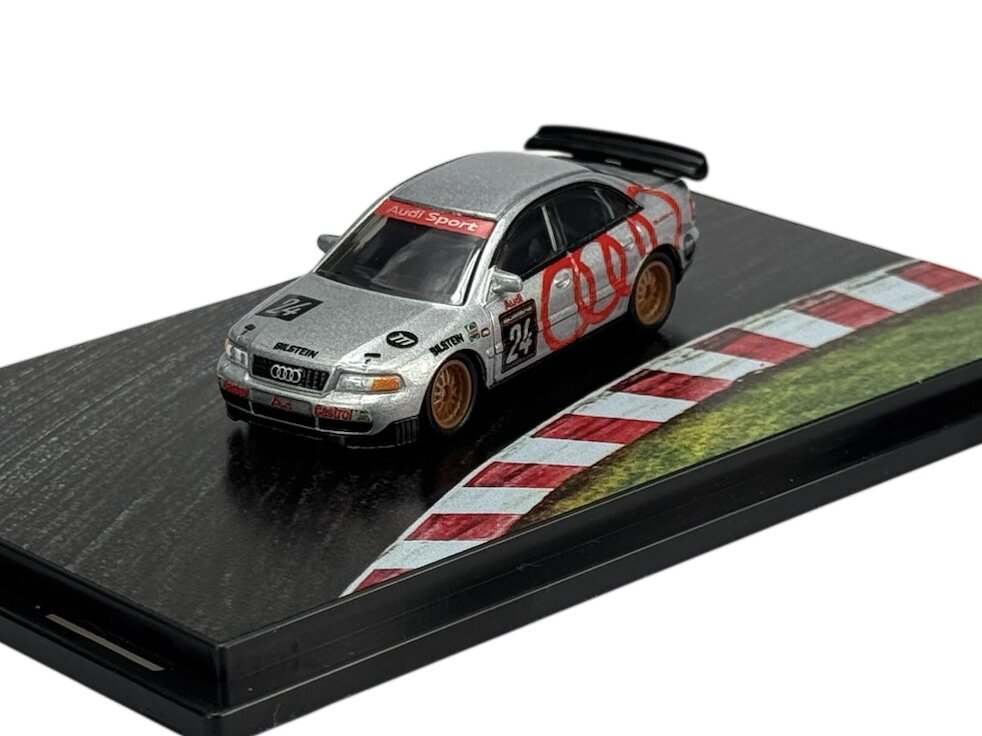 Model samochodu wyścigowego w srebrnym kolorze z numerem 24 na drzwiach i napisem \'Audi Sport\' na przedniej szybie. Samochód stoi na podstawce z grafiką toru wyścigowego.