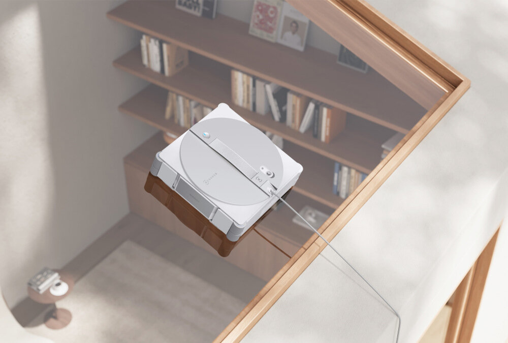 Na szklanej powierzchni znajduje się kwadratowy robot do mycia okien z widocznym logo ECOVACS. W tle widać wnętrze z półkami pełnymi książek.