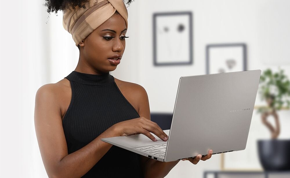 Osoba trzymająca otwarty laptop w jasnym pomieszczeniu z obrazami na ścianie. Na laptopie widoczne jest logo producenta.