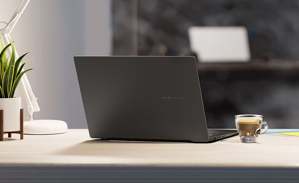 Na biurku stoi zamknięty laptop w ciemnym kolorze, obok którego znajduje się filiżanka z napojem oraz roślina w doniczce. W tle widać rozmyty obraz monitora.