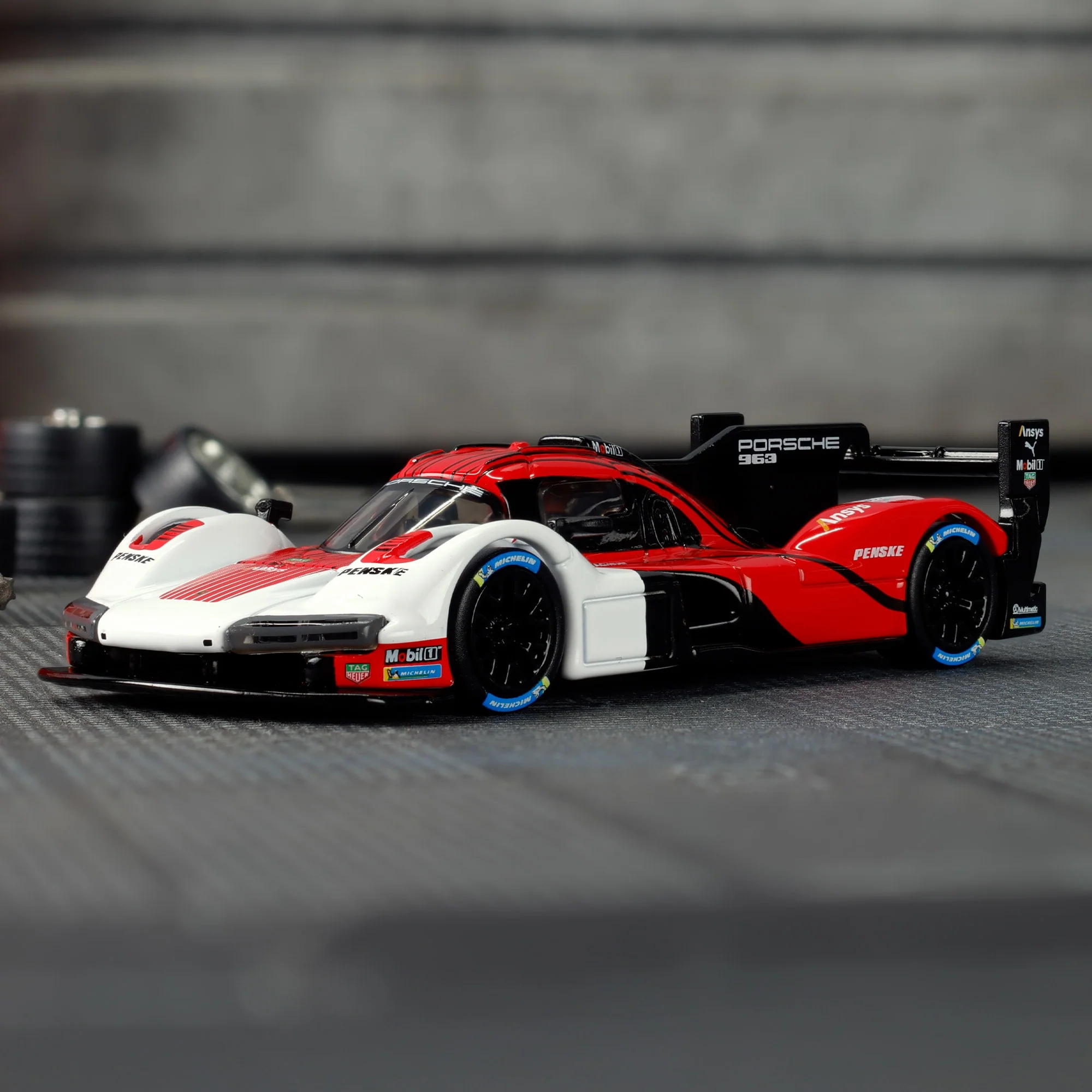 Model samochodu wyścigowego w kolorach białym, czerwonym i czarnym z widocznymi napisami \'Porsche 963\' oraz \'Penske\'. Na oponach znajdują się niebieskie akcenty i logo \'Michelin\'.