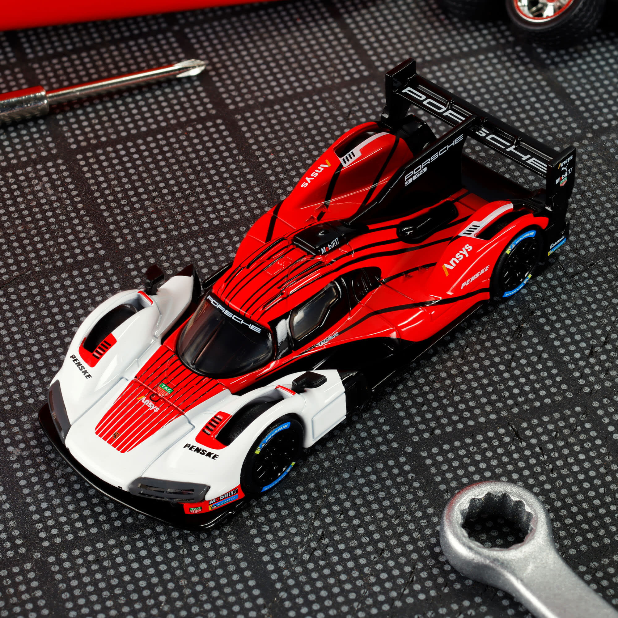 Model samochodu wyścigowego w kolorach czerwonym, białym i czarnym z widocznymi napisami \'Porsche\', \'Penske\' i \'Ansys\'. W tle znajduje się klucz płaski i śrubokręt na kratkowanej powierzchni.