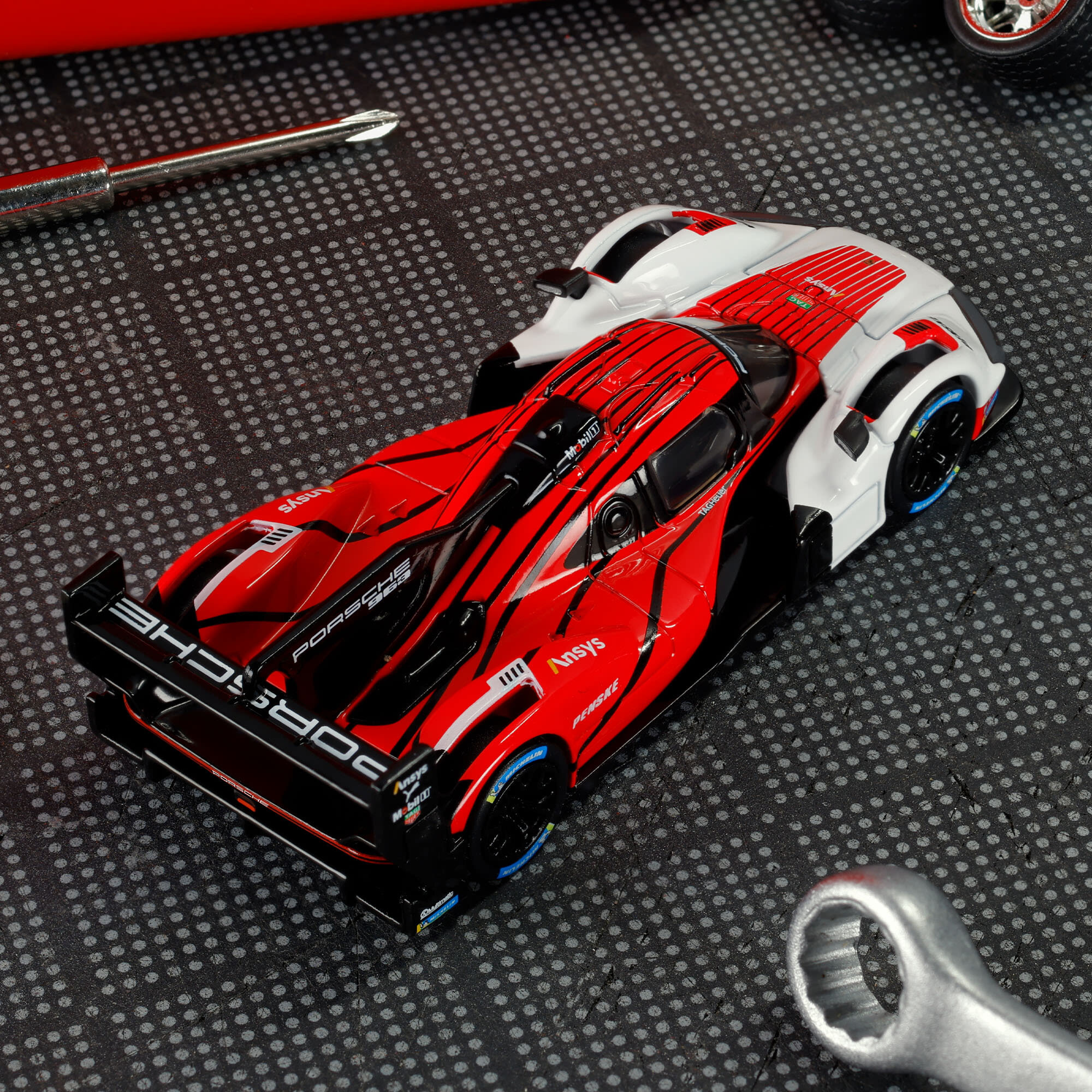 Model samochodu wyścigowego w kolorach czerwonym, czarnym i białym z widocznym napisem \'PORSCHE\' na tylnej części. W tle widoczne są narzędzia na szarej, wzorzystej powierzchni.