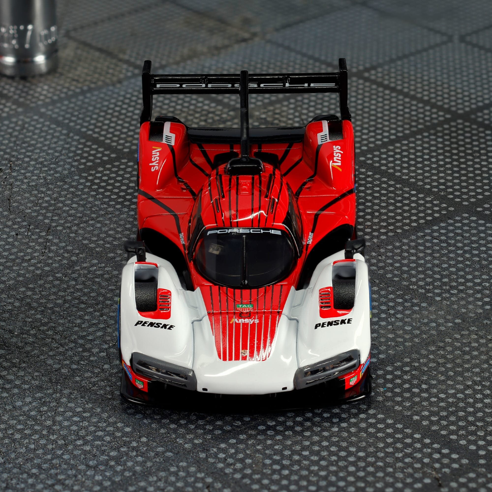 Model samochodu wyścigowego w kolorach czerwonym i białym z widocznymi napisami \'Porsche\' i \'Penske\'. Samochód stoi na powierzchni z wzorem w kratkę.