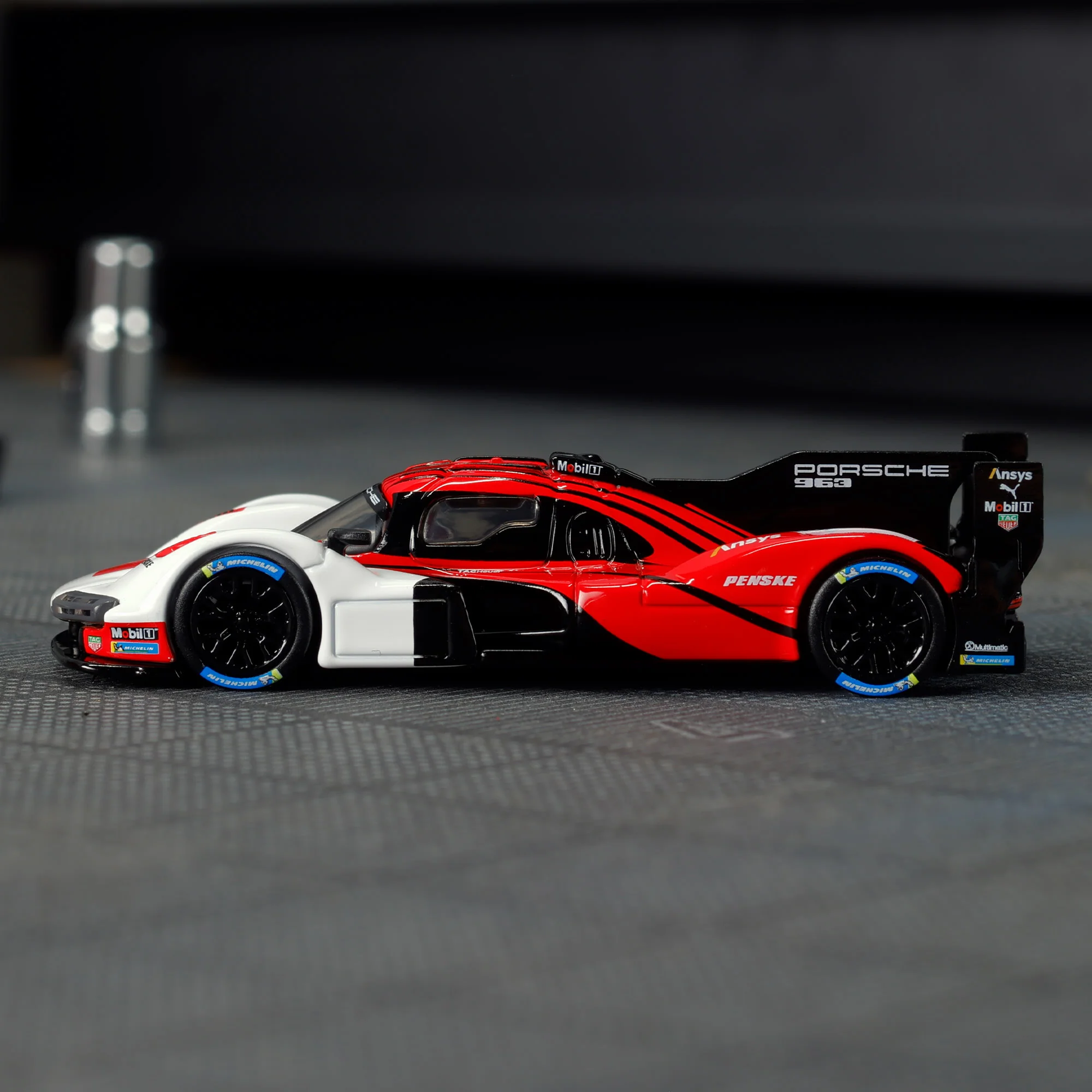Model samochodu wyścigowego w kolorach czerwonym, białym i czarnym z widocznymi napisami \'Porsche 963\', \'Penske\' oraz logo \'Mobil 1\'.