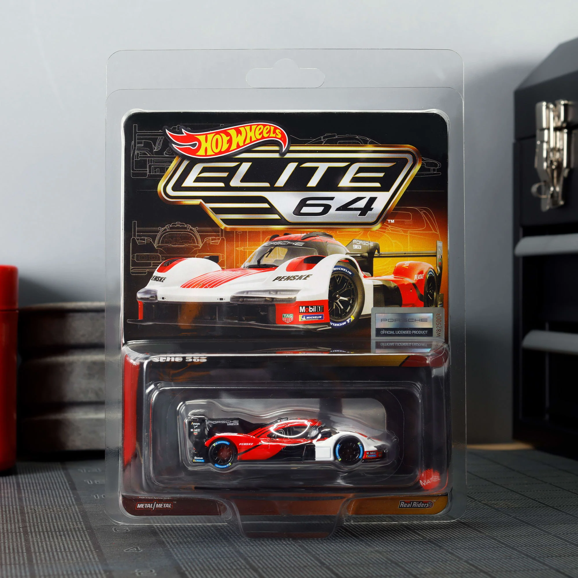 Opakowanie z modelem samochodu wyścigowego Hot Wheels Elite 64, przedstawiające biało-czerwony pojazd z napisem \'Porsche Penske\'. Widoczne logo \'Hot Wheels\' oraz tekst \'Elite 64\'.