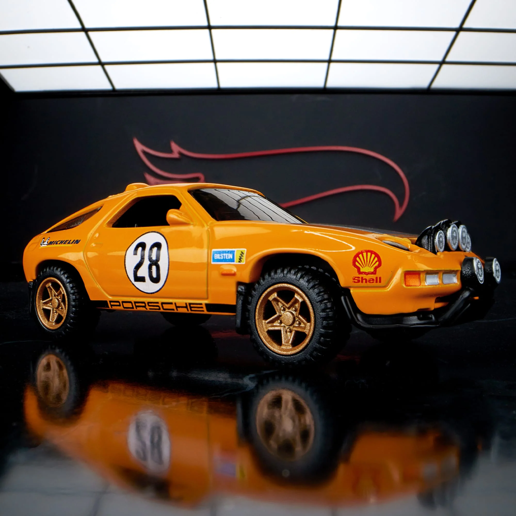 Model samochodu w kolorze pomarańczowym z numerem 28 na drzwiach i napisami \'Porsche\', \'Shell\', \'Michelin\' oraz \'Bilstein\'. Samochód ma złote felgi i dodatkowe reflektory na masce.