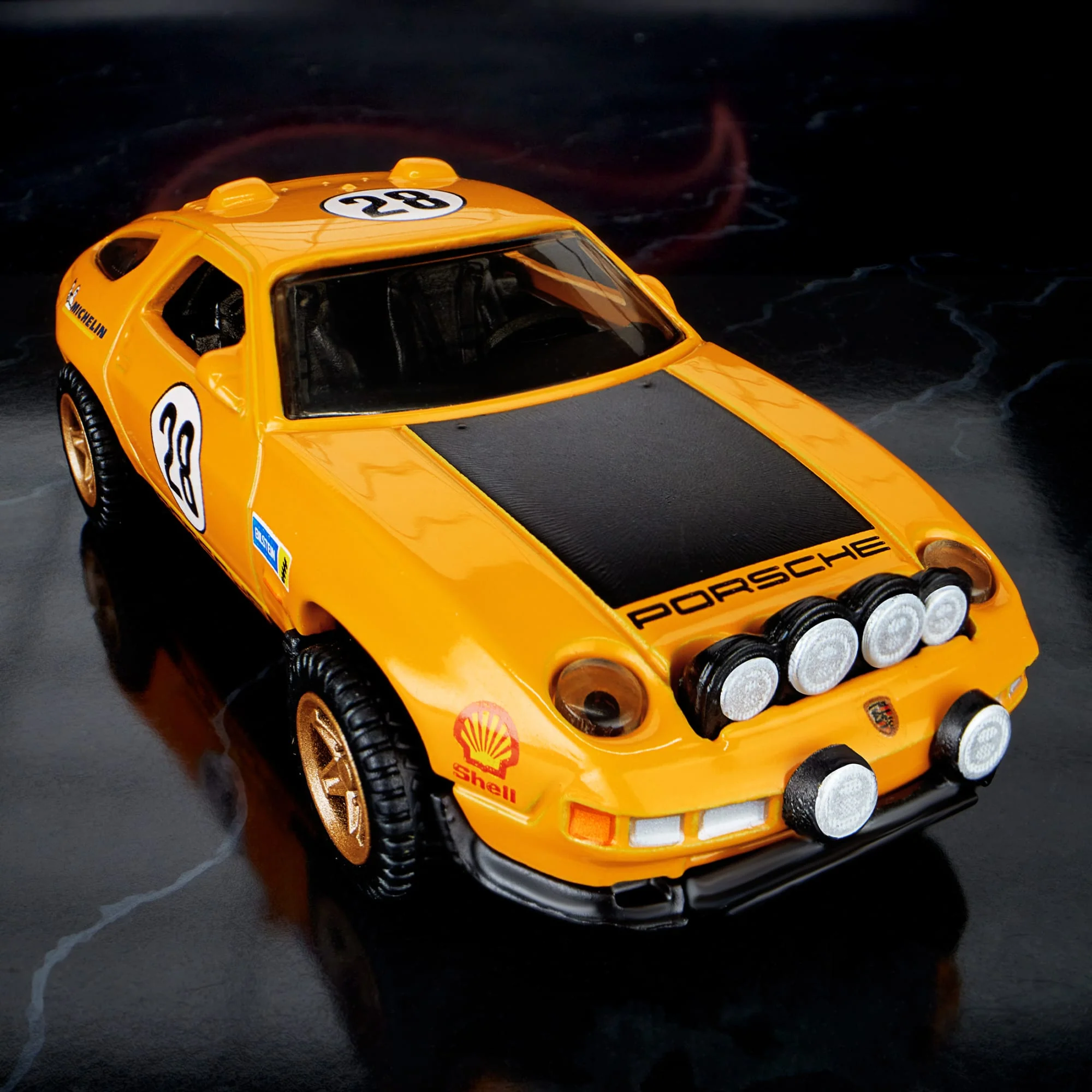 Model samochodu w kolorze pomarańczowym z numerem 28 na masce i drzwiach. Na przodzie widoczne są reflektory oraz logo \'Porsche\' i \'Shell\'.