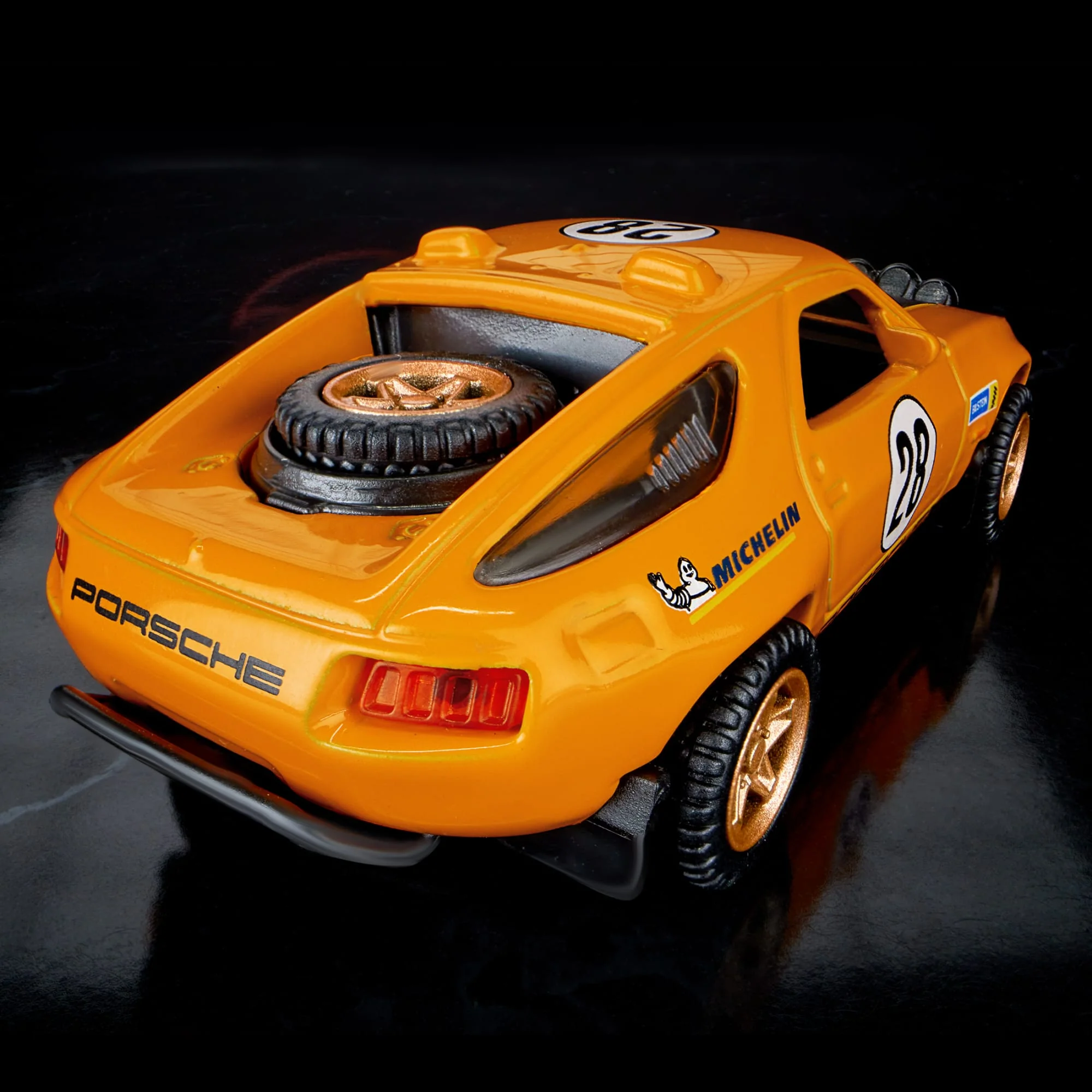 Model samochodu w kolorze pomarańczowym z numerem 28 na drzwiach i logo \'MICHELIN\' na boku. Na tylnej części widoczny jest napis \'PORSCHE\' oraz zapasowe koło.