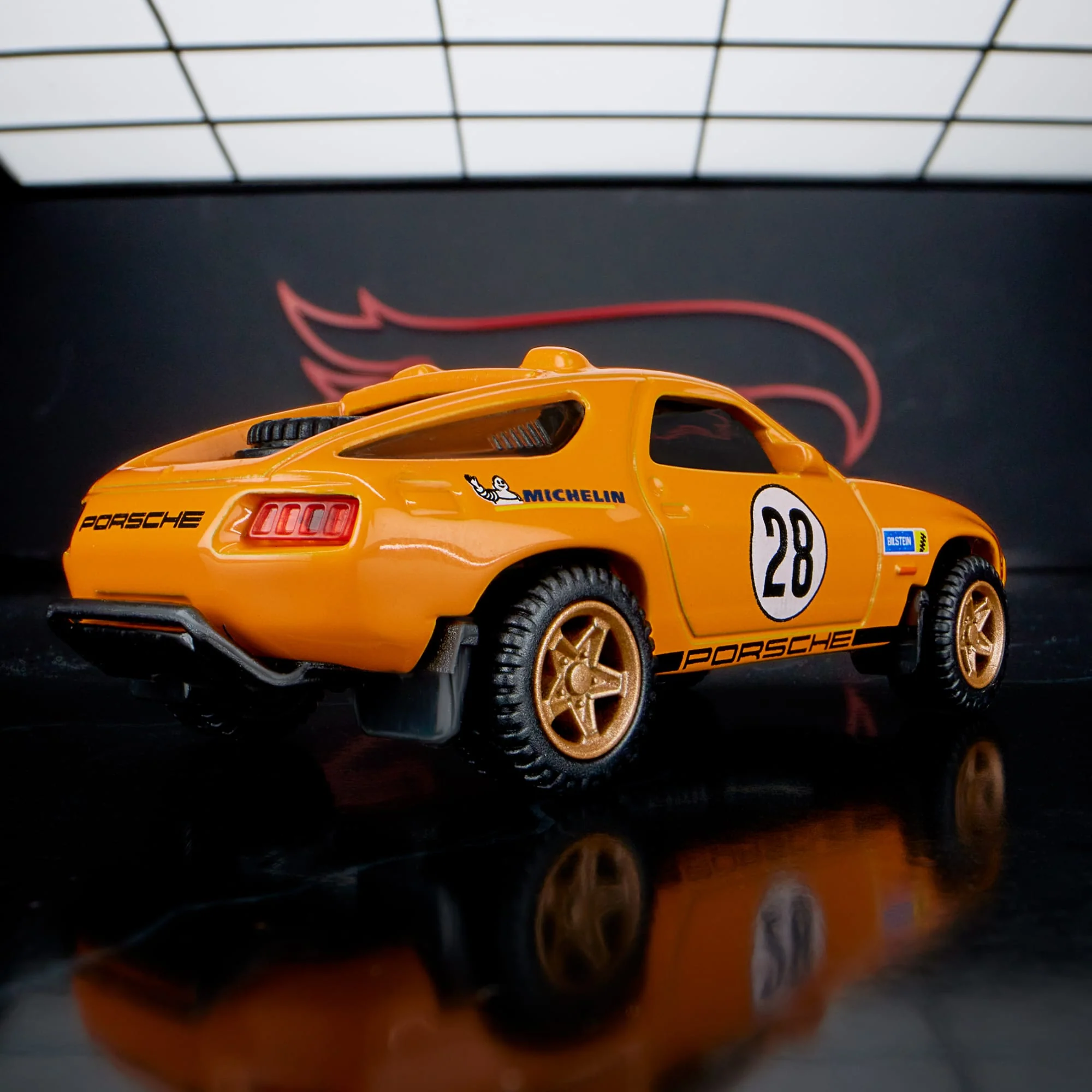 Model samochodu w kolorze pomarańczowym z numerem 28 na boku, oznaczeniami \'Porsche\' i \'Michelin\'. W tle widoczne jest logo w kształcie płomienia.
