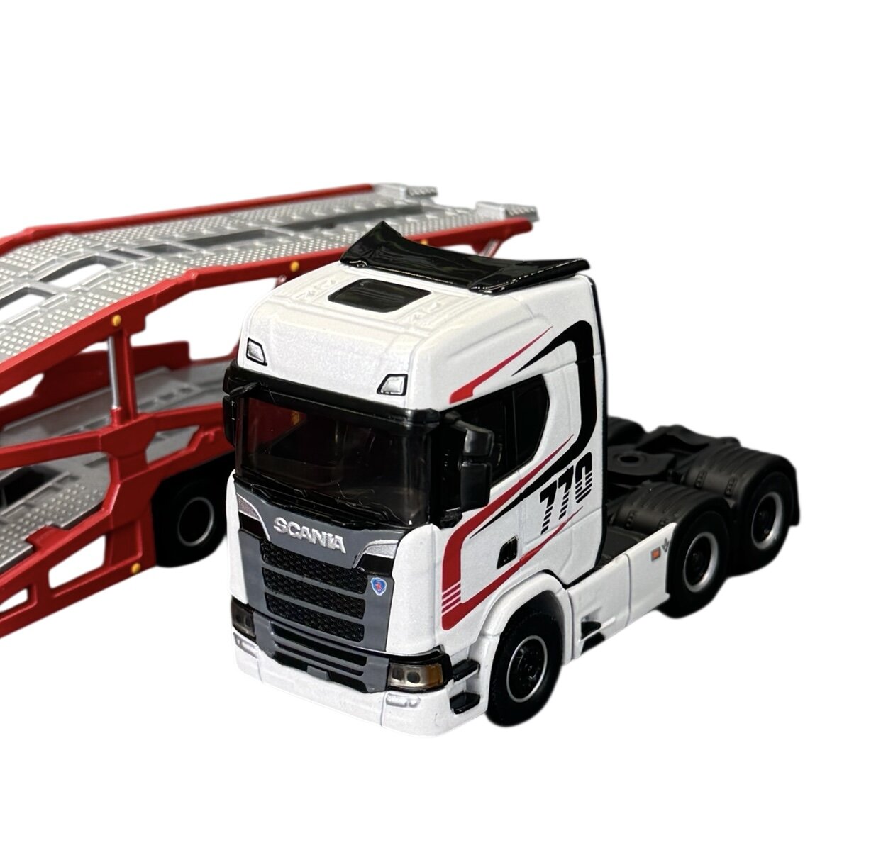 Model ciężarówki w skali 1:64, biały z czarnymi i czerwonymi detalami, z widocznym napisem \'Scania\' i numerem \'770\'. W tle znajduje się czerwono-srebrna naczepa.